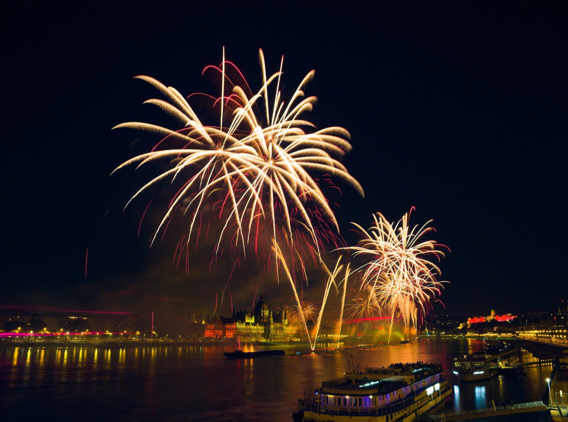 fireworks over the danube in budapest, seen from the margidhíd (margaret bridge), ships in the foreground, parliament in the middle, behind the fireworks, and the castle to the right